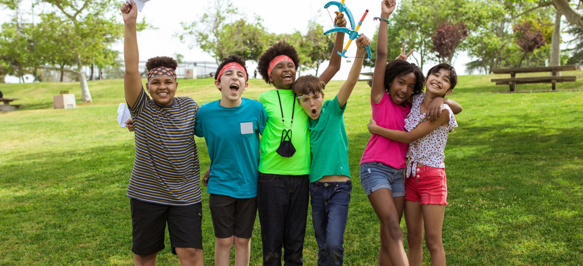 A cheerful group of children playing and celebrating with arms raised at a park on a sunny day.