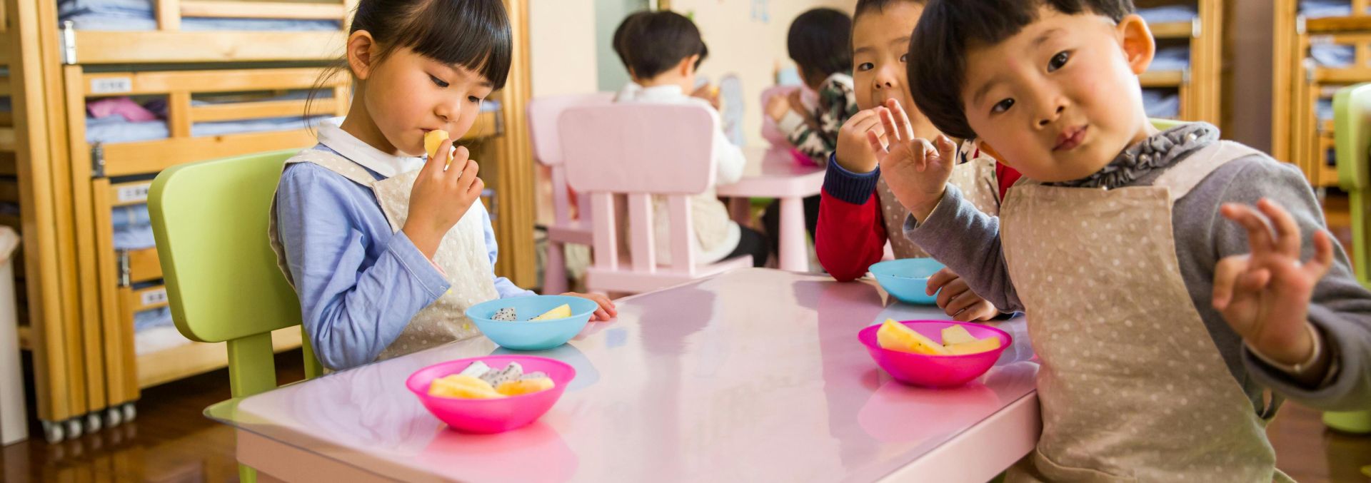 Kids seated around a table in a colorful classroom, eating snacks happily.