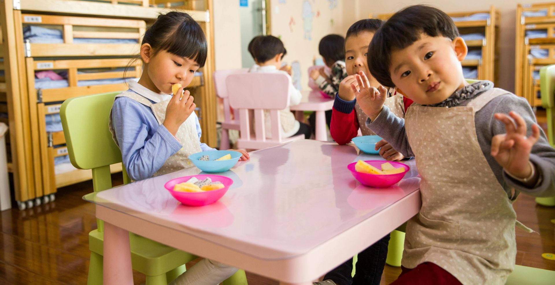 Kids seated around a table in a colorful classroom, eating snacks happily.