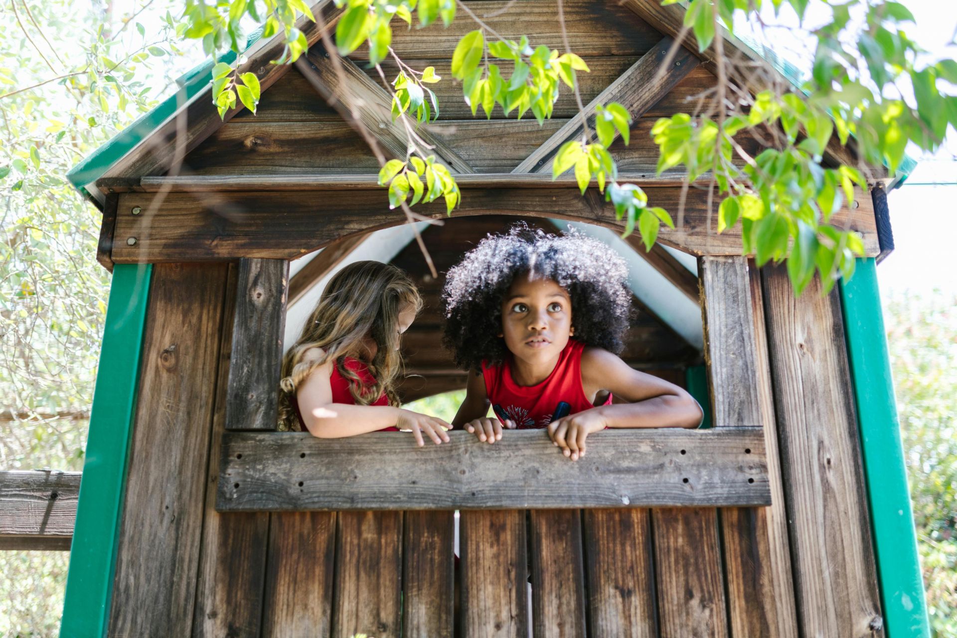 Two children exploring and playing in a wooden treehouse surrounded by green leaves.