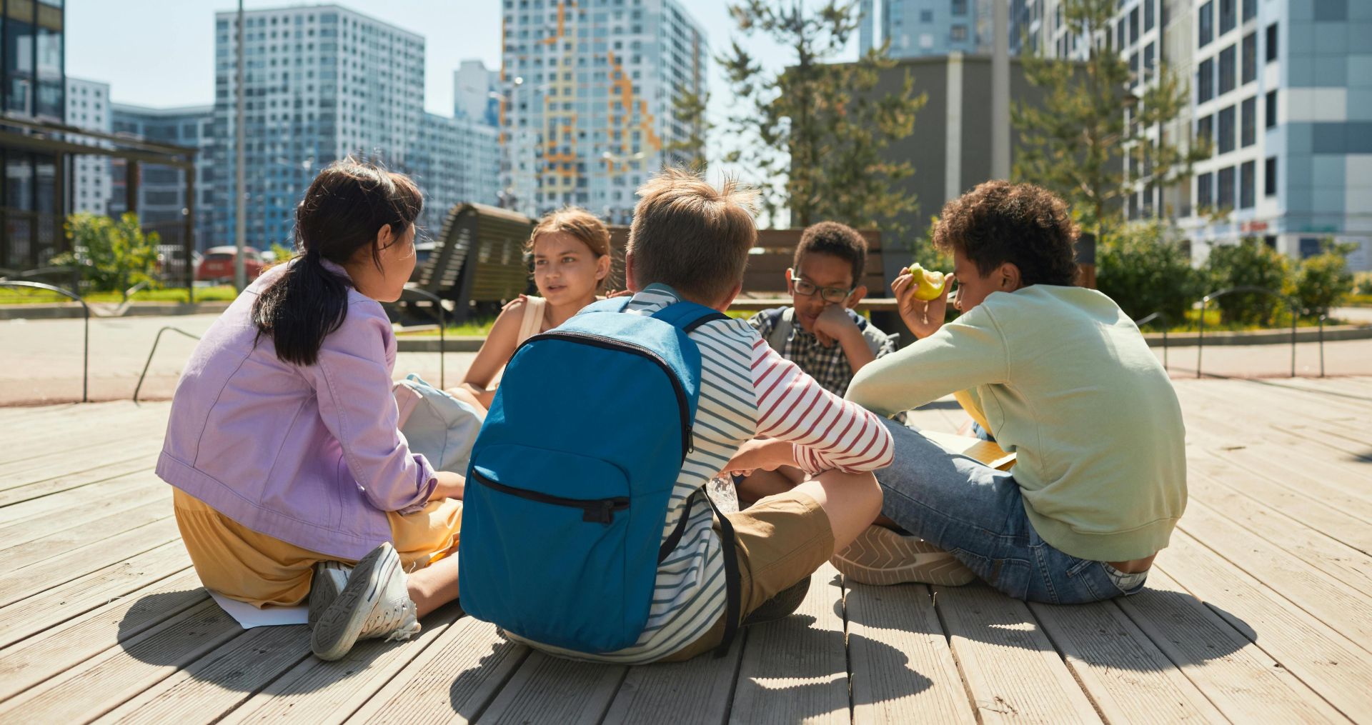 A diverse group of children sits together enjoying lunch on a sunny day in the city.