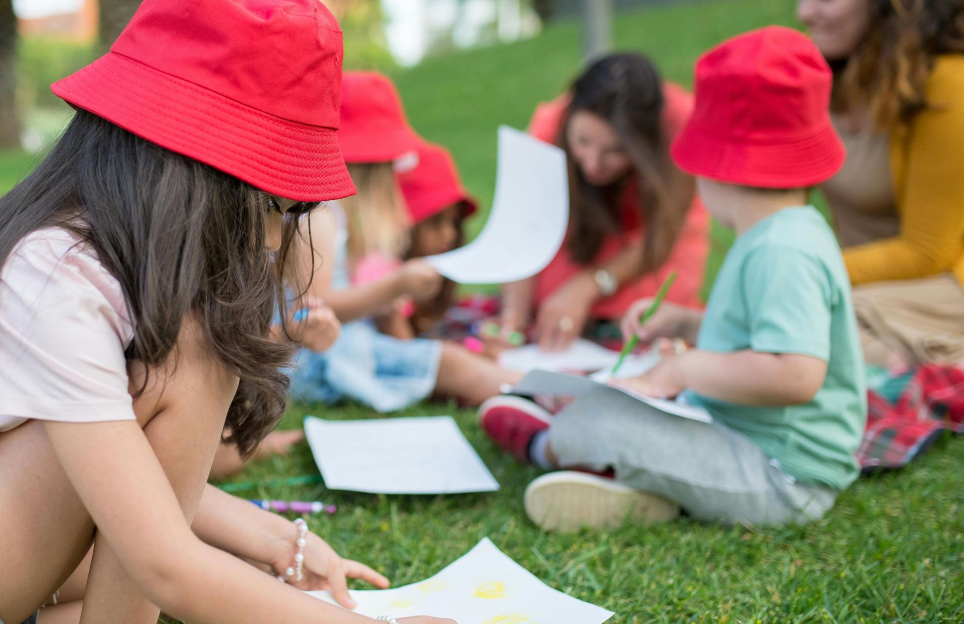 Children engaged in outdoor drawing activities on a sunny day wearing red bucket hats.