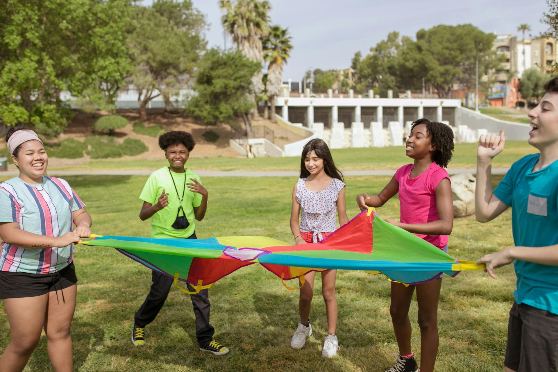 A joyful group of kids playing with colorful parachute in a green park under sunny skies.